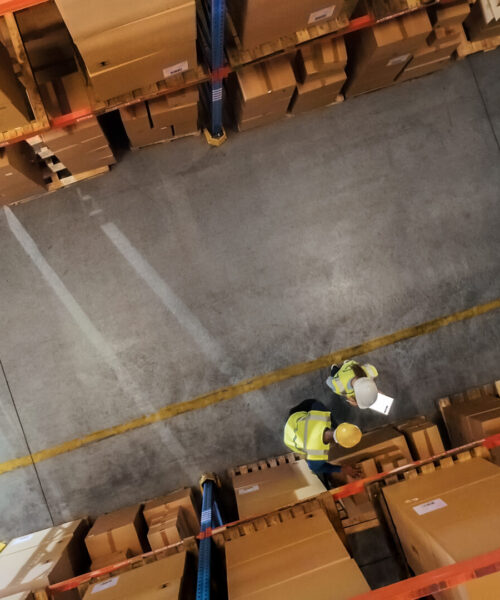 Top-Down View: Workers Stand in Rows of Shelves with Goods in Retail Warehouse, Use Digital Tablet and Talk About Package Delivery. People Work in Product Distribution Logistics Center