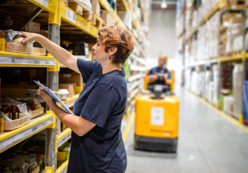 Woman with checklist picking jar from package on warehouse rack