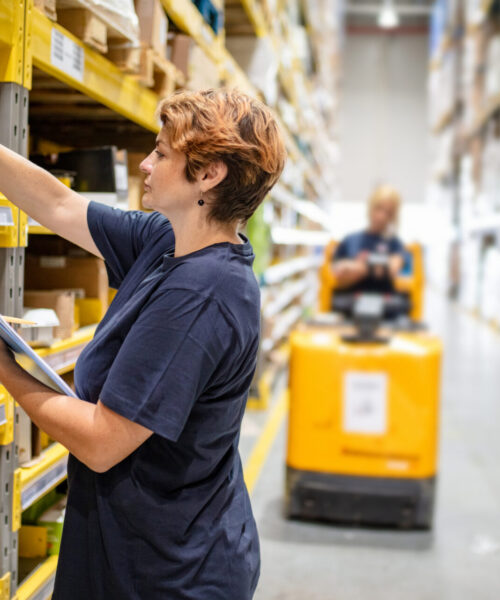 Woman with checklist picking jar from package on warehouse rack