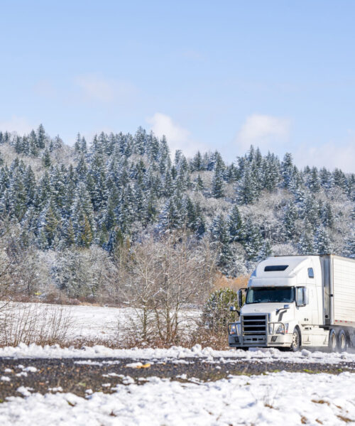 Big rig long haul white semi truck tractor transporting commercial cargo in refrigerator semi trailer going on the wet glossy road with water from melting snow and winter snowy trees on the hill
